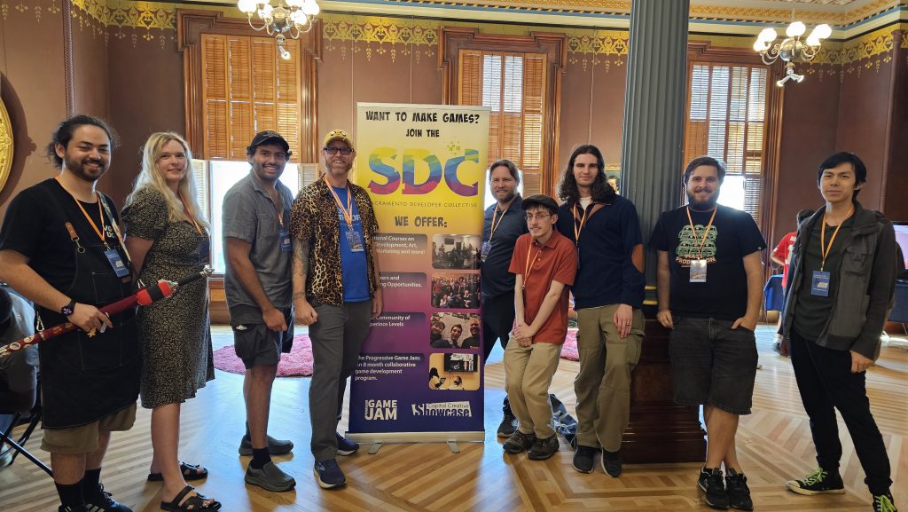 A group of ten people wearing event badges pose around a colorful “Join the SDC” banner inside an ornate room with tall windows, wood floors, and chandeliers.